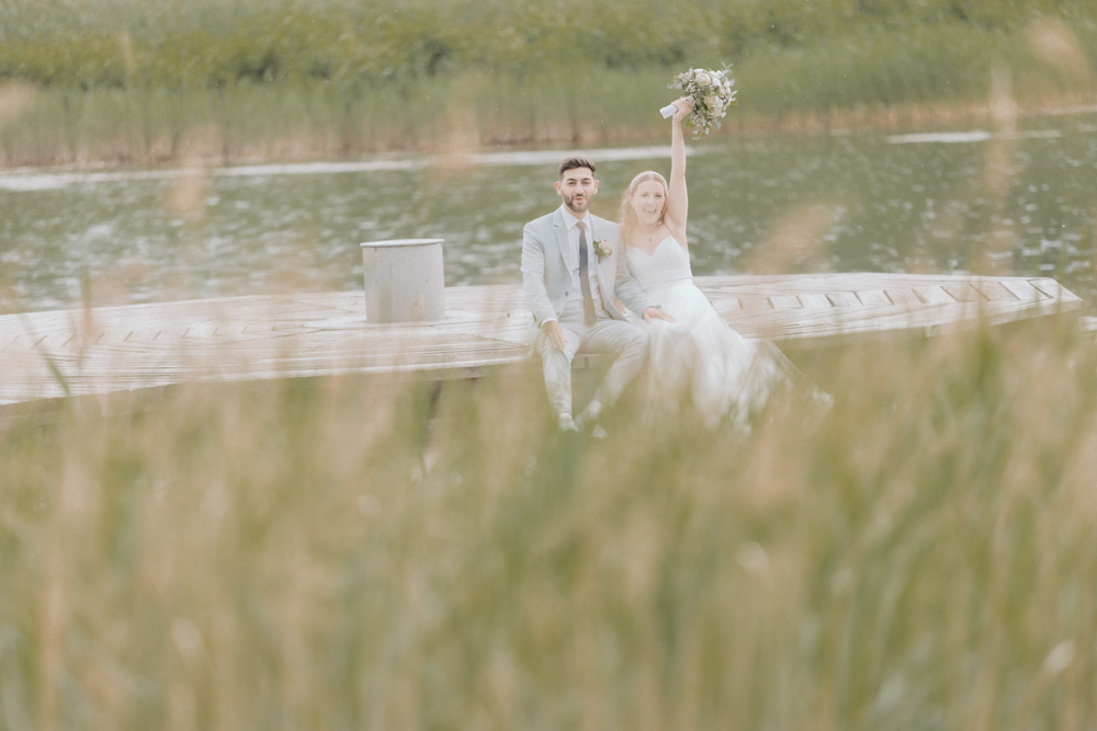 Brautpaarshooting im Britzer Garten am See. Es scheint die Sonne und das Brautpaar sitzt auf der Holzplattform. Die Braut hält ihren Brautstrauß in die Luft. Das Brautpaarshooting ist Teil der Hochzeitsreportage und des Hochzeitsvideos.