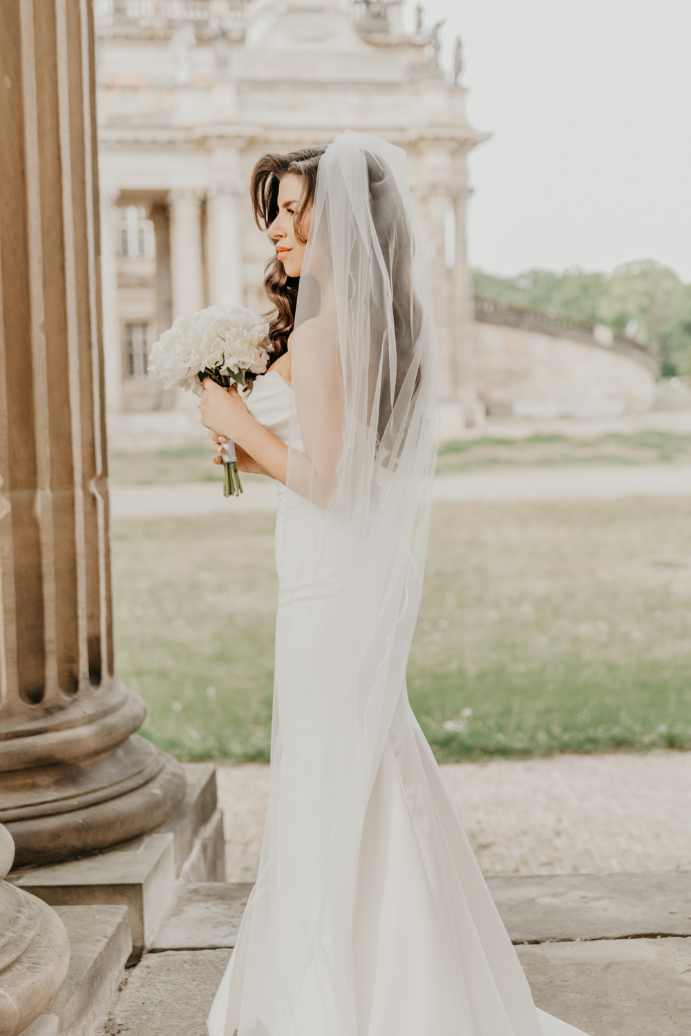Hier wird die Braut alleine, im Rahmen des Brautpaarshootings fotografiert. Das Shooting findet am Neuen Palais in Potsdam statt. Die Braut steht alleine im Bogen vor einer Sandsteinsäule und hält ihren Brautstrauß in den Händen. Teil der ganztägigen Hochzeitsreportage.