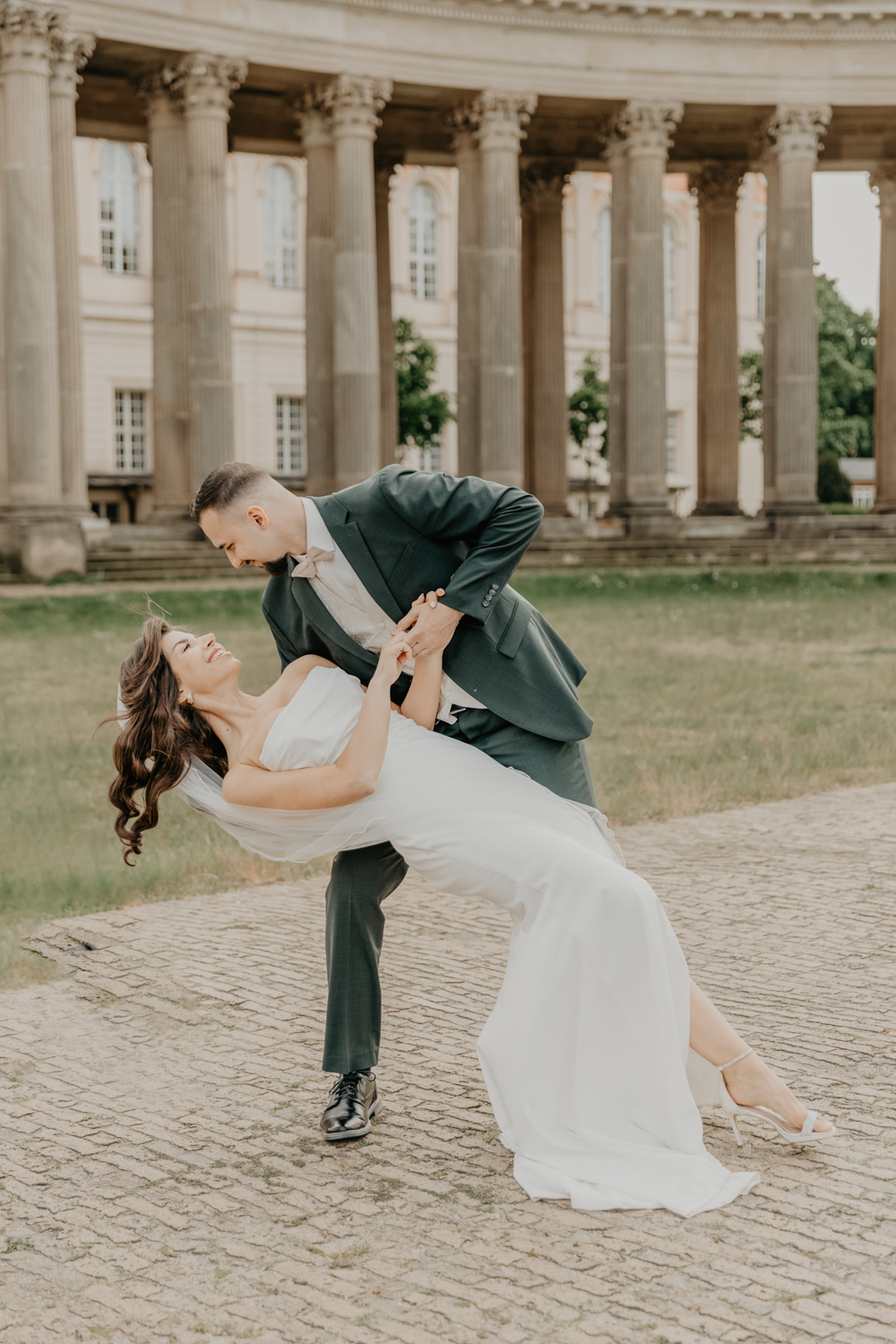 Der Bräutigam schaut seine Braut an. Das Brautpaar tanzt gerade im Park Sanssouci. Der Bräutigam haält seine Frau fest an den Händen. Die Braut lehnt sich nach hinten. Das Shooting fand am Neuen Palais im Schloßpark Sanssouci in Potsdam statt und war Teil einer ganztägigen Hochzeitsreportage.