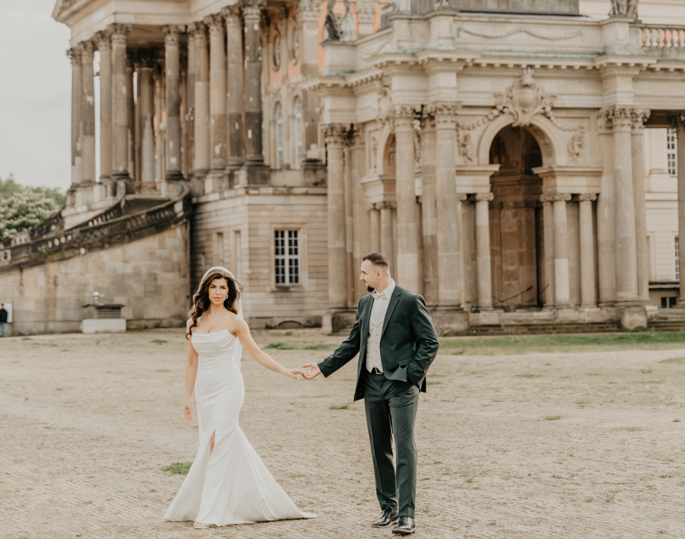 Das Brautpaar spaziert auf dem großen Platz vor einem alten Gebäude. Das Brautpaarshooting war Teil der ganztätigen Hochzeitsreportage. Das Shooting mit dem Brautpaar fand am Neuen Palais in Potsdam statt.
