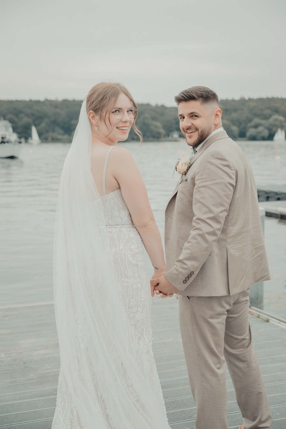 Das Brautpaar steht am See auf einem Steg. Sie schauen dabei in die Kamera und halten sich an den Händen. Das Foto entstand im Rahmen einer Hochzeitsreportage in Berlin.